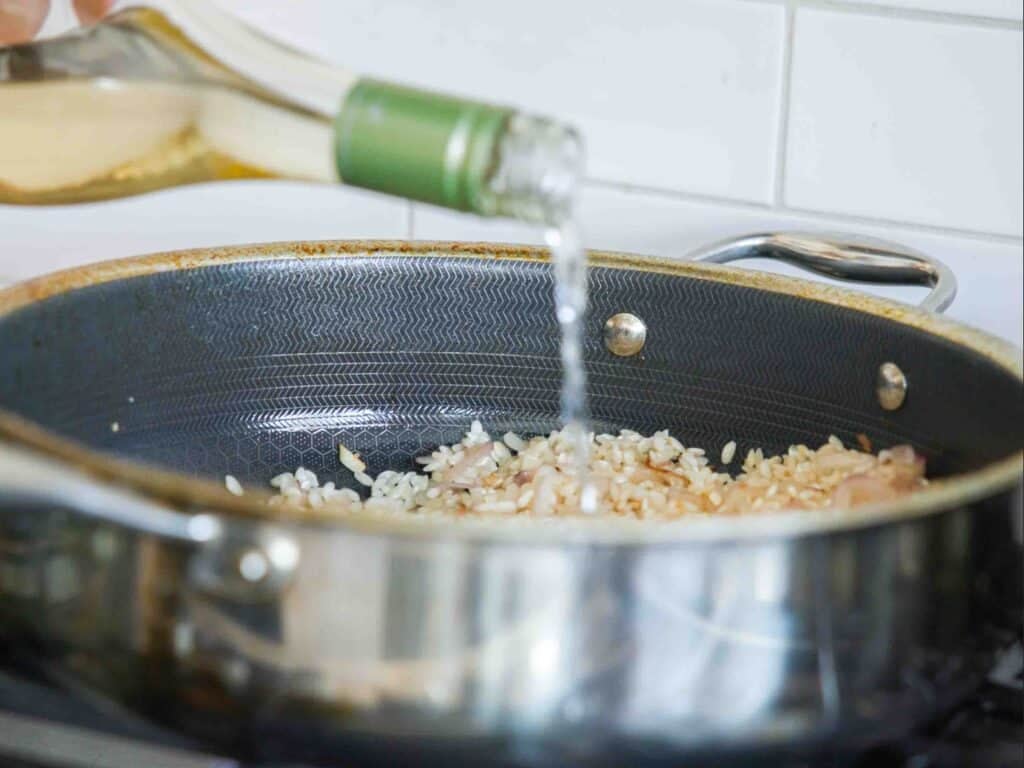 A close-up of a pan on a stove with chopped onions and rice being cooked for green pea risotto, while white wine is being poured in from a bottle.